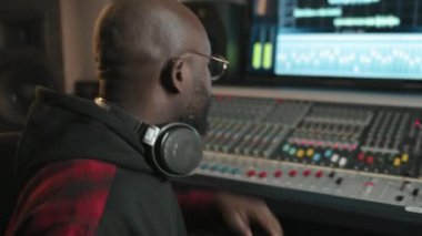 Gözlüklü Afrikalı Afrikalı Amerikalı erkek prodüktör müzik dinliyor ve stüdyosundaki iş yerinde otururken kayıt panosundaki konsolları dengeleyerek sesi ayarlıyor.