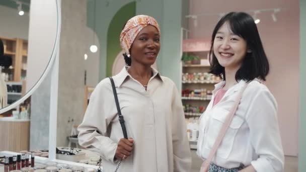 モダンなスーパーのカメラの前に立つ白いカジュアルなシャツの2人の幸せな若い多文化女性ディスプレイ上の化粧品の様々なと笑顔であなたを見て — ストック動画
