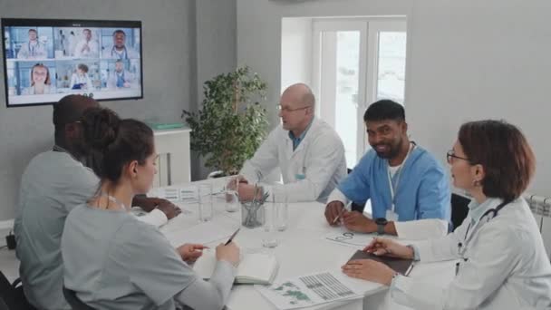 Grupa Wieloetnicznych Lekarzy Siedzących Przy Stole Omawiająca Kwestie Współpracownikami Podczas — Wideo stockowe