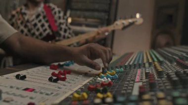 Genç Afrikalı Amerikalı yapımcının eli, yeni bir müzik parçası işlerken ve elektro gitar çalan müzisyene karşı kayıt yaparken konsolları ses karıştırıcı üzerinde dengeliyor.