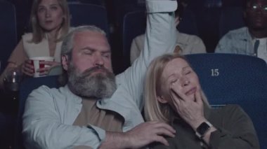 Yetişkin, beyaz, sakallı bir adam sinemada acıklı dram filmi izlerken karısına sarılıyor.
