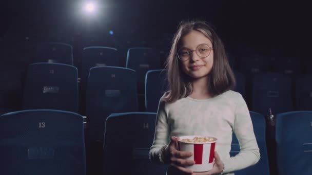 身着眼镜 拿着爆米花桶 站在电影院里 靠着海军蓝色座椅的白人少女的中等长度肖像 — 图库视频影像