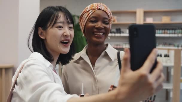 在化妆品超级市场的智能手机里 年轻的亚洲女人在涂上新的唇彩后 用智能手机看了看自己的形象 然后与非洲裔美国朋友自拍 — 图库视频影像