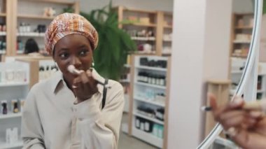 Afro-Amerikalı bir kız kozmetik süpermarketinde aynanın önünde dikilirken gülümseyerek kameraya bakıyor ve yeni barutu test ediyor.