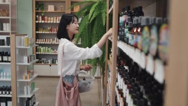 化粧品スーパーで巨大なディスプレイの前に立って肩にハンドバッグとアジアの女の子 ボディケア製品を選択し ショッピングバスケットにそれらを入れて — ストック動画