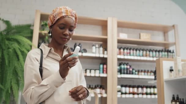 スキンケア製品を持つ小さなボトルを見て 化粧品スーパーで新しいセルフケアアイテムを選択しながら買い物かごに入れる若い黒人女性 — ストック動画