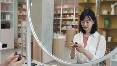 Genç Asyalı bir kadın kozmetik süpermarketinde farklı renkler denerken aynanın karşısında dudaklarına kırmızı ruj sürüyor.