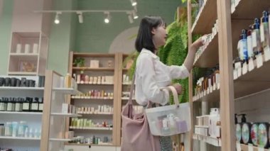 Genç Asyalı alışverişkolik, kozmetik ürünleri raftan alıyor, alışveriş sepetine koyuyor ve geniş bir süpermarkette güzellik malzemeleri satan başka bir sergiye gidiyor.