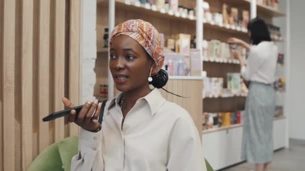 年轻的非洲裔美国女买家坐在化妆品超级市场的相机前 一边用智能手机记录语音信息 一边与其他消费者谈论选购美容产品的背景 — 图库视频影像