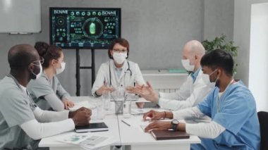 Maske takan farklı etnik gruplardan oluşan bir grup doktor masaya oturuyor, olgun pratisyen hekimler karantina sorunları hakkında konuşuyor.