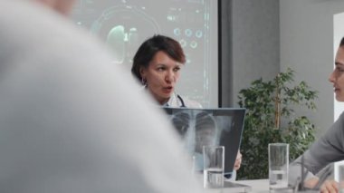 Yavaş çekim orta ölçekli, göğüs röntgeni çeken olgun bir kadın doktor. Meslektaşlarıyla aynı masaya oturmuş teşhis koyuyor.
