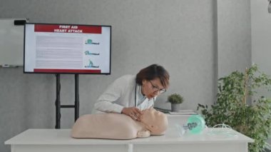 Profesyonel kadın sağlık görevlisinin ilk yardım ve kalp masajını nasıl yapacağını açıklayan bir kuklayla masanın önünde durduğu orta boy bir fotoğraf.