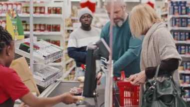 Kadın veznedarla süpermarkete giriş yapan müşteriler kuyruğunda oturup ürünlerini tarıyorlar.