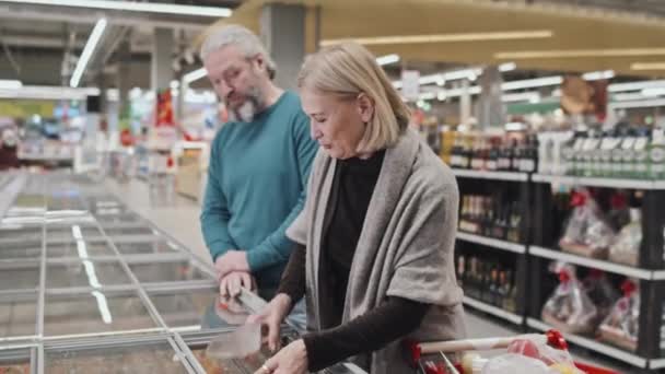 Зрелая Белая Пара Стоящая Морозилки Супермаркете Выбирает Покупает Смешанные Овощи — стоковое видео