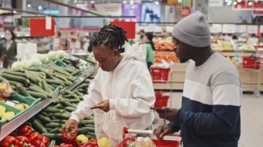 Süpermarketten taze ürün almak için alışveriş yapan genç Afrikalı Amerikalı çift, sebzeleri tekrar kullanılabilir ürün torbalarına koyuyorlar.