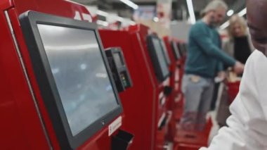 Genç Afrikalı Amerikalı kadın süpermarkette kendi kendini kontrol eden makinenin yanında duruyor ve satın alınan ürünlerin parasını kredi kartıyla ödüyor.