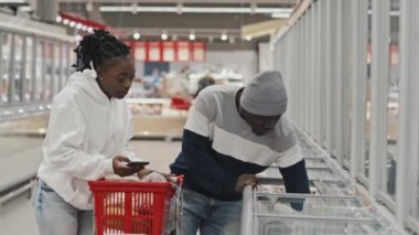 Genç Afrikalı Amerikalı çift büyük süpermarkette yiyecek alırken dondurulmuş ürünler alıyor.