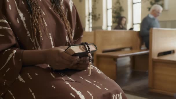 選択的焦点中セクションショットの認識できない黒の女性保持祈りの本とロザリオ座っている上のベンチで教会 — ストック動画