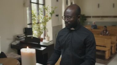Kilisede mihrabın önünde durup dua eden ve haç çıkaran Afrikalı Amerikalı Katolik rahibin orta boy portresi.