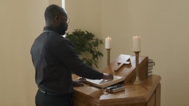 カトリック教会の木製の祭壇に立つ若い大人のアフリカ系アメリカ人司祭の中のショット聖書を読む — ストック動画