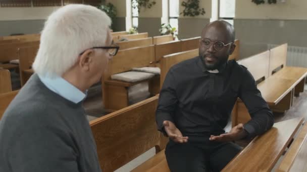 高角度ショットの若いです大人アフリカ系アメリカ人カトリック聖職者座ってベンチに教会で話してシニア白人教区 — ストック動画