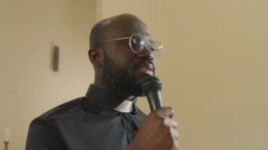 Kilise ayini sırasında mikrofonla konuşan genç Afrikalı Amerikalı Katolik rahibin orta ölçekli düşük açılı görüntüsü.