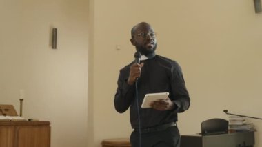 Elinde dijital tablet ve mikrofon tutan Afrikalı Katolik rahibin kilise cemaatine vaaz verirken orta açılı portresi.