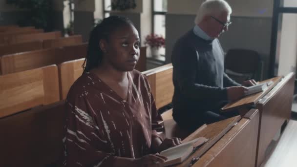 高角度ズームで現代の若いアフリカ系アメリカ人女性のベンチにカトリック教会で牧師に耳を傾け 聖書を読む — ストック動画
