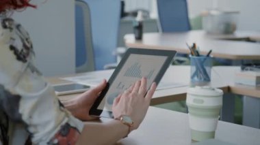 Ofiste masasında oturan tanınmayan bir kadın dijital tablet ekrandaki grafik ve çizelgelere bakıyor.