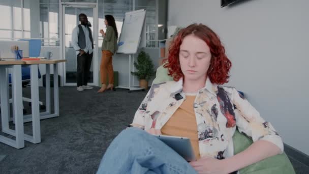 在现代写字楼里 穿着休闲装坐在豆袋椅子上做数码平板电脑的红卷女青年的肖像 — 图库视频影像