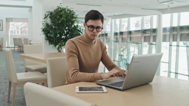 Bilgisayarda yazarken dijital tablet bilgilerini kontrol eden ciddi genç iş adamının orta boy görüntüsü.