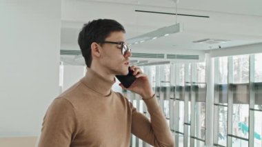 Gözlüklü genç işadamının modern ofiste dikilirken cep telefonuyla konuştuğu orta ölçekli yakın çekim.