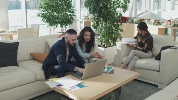 オフィスホールでソファに座っている若いビジネスカップルの広いショットは ラップトップ上でオンラインプレゼンテーションで作業 — ストック動画