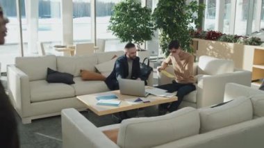 Ofisteki kanepede otururken iki genç işadamı belgelere bakıyor ve bilgisayarlarını kullanıyorlar.