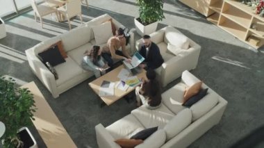 Bir grup iş adamının masanın etrafında oturup ofis salonundaki projeyi tartıştığı yüksek açılı bir manzara.