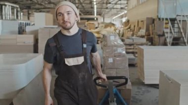 Mobilya fabrikasında çalışırken karton kutularla el arabası çeken genç bir adam.