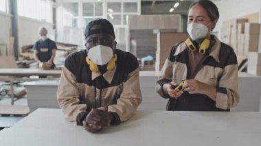Beyaz kadın işçi koruyucu kulaklıklar ve maskeyle Afrikalı Amerikalı meslektaşına yürüyor ve mobilya fabrikasında fotoğraf makinesi için birlikte poz veriyorlar.
