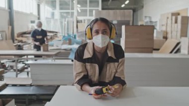 Koruyucu kulaklık ve maske takan, tahta tahtaya yaslanan ve mobilya fabrikasında kamera önünde poz veren kadın işçinin portresi.