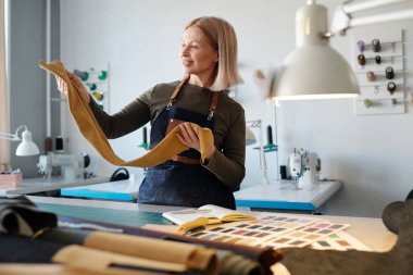 İş kıyafetleri içindeki mutlu sarışın kadın, iş yerinde iş malzemeleriyle ayakta dururken elindeki süet ya da deri parçasına bakıyor.