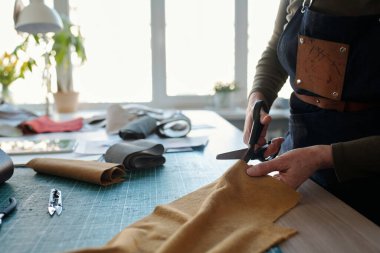 Close-up of female leatherworker in apron cutting leather or suede while standing by table in front of camera and creating new attire clipart