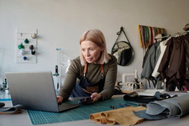 Deri iş yerinin olgun sarışın sahibi iş yerinde dizüstü bilgisayara bakıyor ve yeni online siparişleri inceliyor.