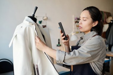 Genç esmer terzi ya da moda tasarımcısı müşteriye göndermeden önce yeni beyaz deri ceketin fotoğrafını çekiyor.