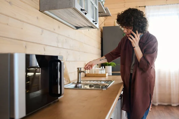 年轻的家庭主妇拿着智能手机 一边站在柜台旁一边 一边打电话给修理工 一边在厨房里用水槽和水龙头敲了一下 — 图库照片