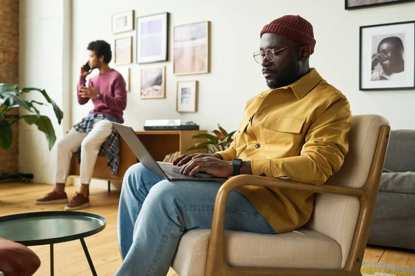 年轻而严肃的非洲裔美国分析家坐在扶手椅上与男性同事在智能手机上交谈时 在笔记本电脑键盘上打字 — 图库照片