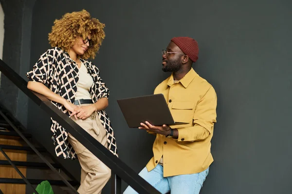 两名穿着便服站在楼梯上讨论在线数据的年轻非洲裔美国同事 其中一人在使用笔记本电脑 — 图库照片