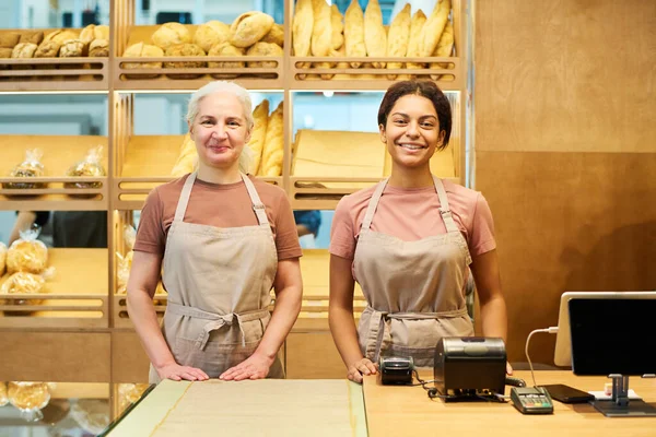 パン屋のカウンターデスクで制服姿の2人の陽気な女店員と新鮮なパンでディスプレイに対するカメラを見て — ストック写真
