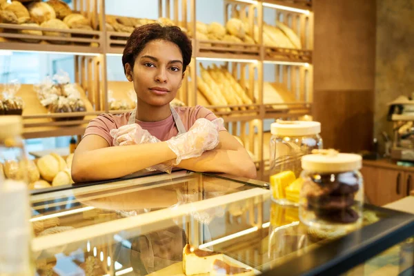 若いですかわいい女性店員立ってガラスディスプレイでペストリーでパン屋さんと新鮮なパンと棚に対するカメラを見て — ストック写真