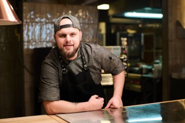 Kendine güvenen genç bir aşçı ya da üniformalı bir aşçı modern restoranın mutfağında duruyor ve kameraya bakıyor.