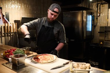 Pizzacının mutfağında çalışırken pizzanın üzerine malzeme döken üniformalı genç bir aşçı.