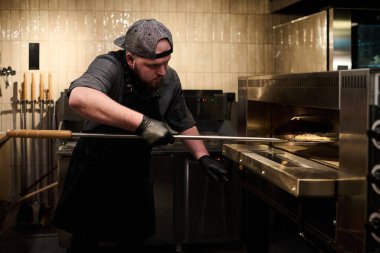 Üniformalı genç aşçı pizzacı mutfağında ekipmanların önünde dururken fırına pizza koyuyor.
